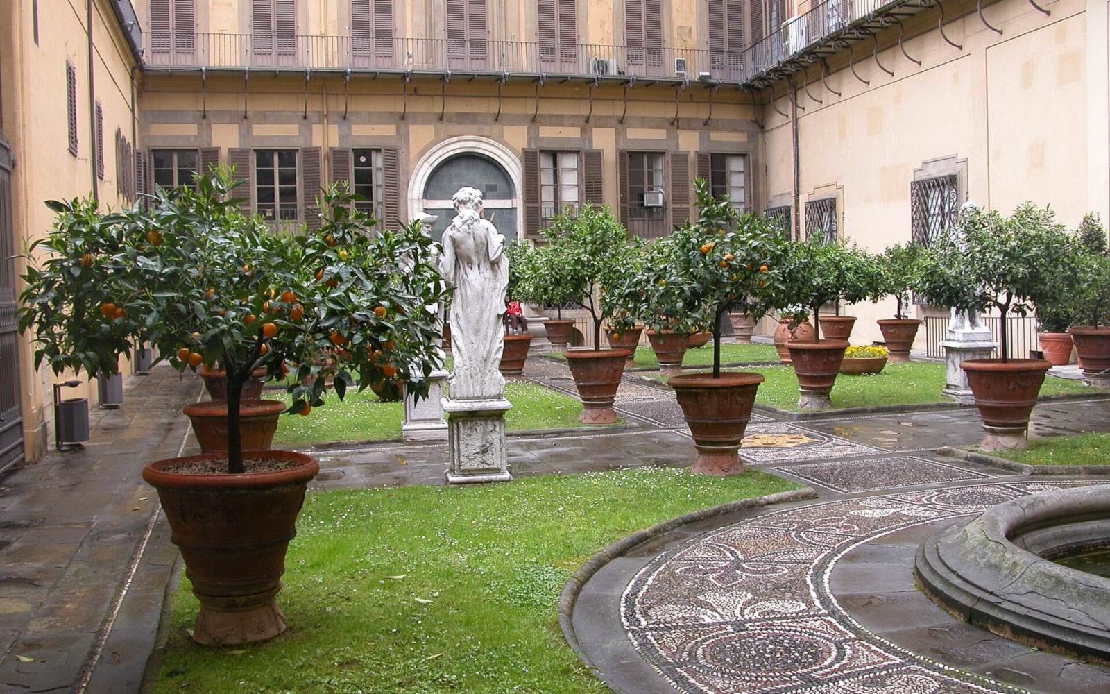 A shaded inner courtyard planted with citrus trees in terracotta pots, lawn and patterned stone paving between building facades.